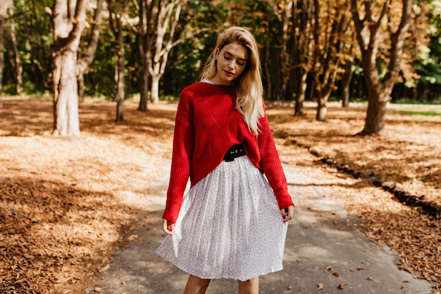 Urocza blondynka pozowanie w parku jesienią. Ubrana w piękną białą sukienkę i ładny czerwony sweter.