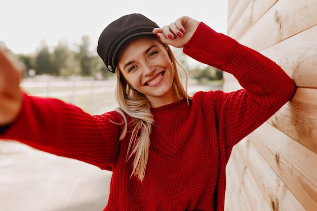 Urocza blondynka w czerwonym swetrze i czarnym kapeluszu radośnie uśmiecha się do swojego telefonu Piękna kobieta robi selfie w pobliżu drewnianego domu w parku