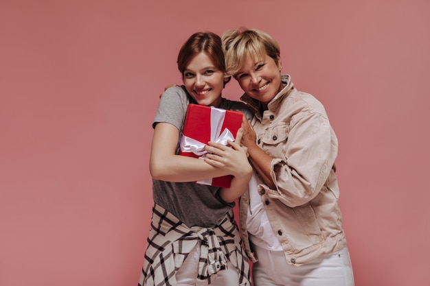 Urocza Brunetka Dama W Szarej Koszulce I Koszuli W Kratę, Trzymając Czerwone Pudełko, Uśmiechając Się I Pozując Ze Starą Blondynką Na Na Białym Tle.