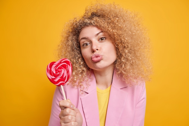 Bezpłatne zdjęcie urocza czuła kobieta z kręconymi włosami trzyma złożone usta trzyma pyszny cukierek na patyku ma słodycze ubrana w formalne ubrania izolowane nad żółtą ścianą wysyła pocałunek w powietrzu