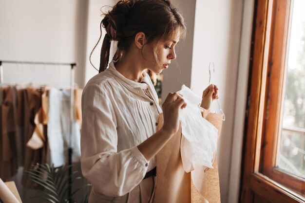 Urocza dama patrząca na swoje zaprojektowane ubrania Atrakcyjna młoda kobieta w białej bluzce pracuje nad swoją nową kolekcją mody