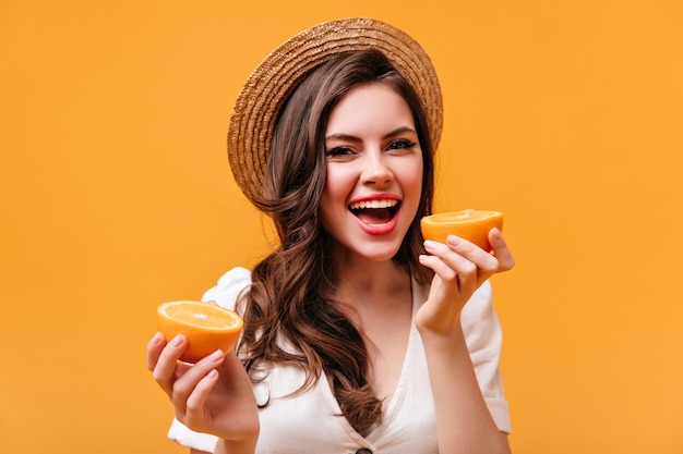 Urocza dama w białej koszulce i słomkowym kapeluszu patrzy w kamerę z uśmiechem i trzyma plasterki pomarańczy.