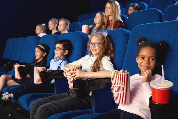 Urocza dziewczyna siedzi w kinie z przyjaciółmi, patrząc na kamery i uśmiechając się podczas oglądania filmu. Małe urocze afrykańskie dziecko kobiece jedzenie popcornu i picie słodkiej wody