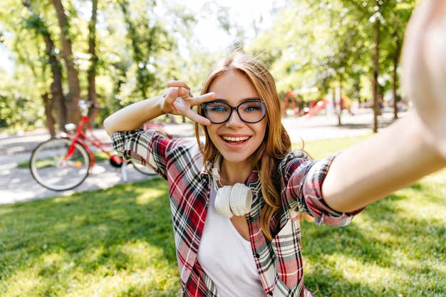 Urocza dziewczyna w okularach co selfie w parku. Plenerowe ujęcie niesamowitej blondynki noszącej słuchawki w letni dzień.