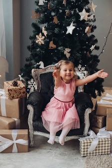 Urocza dziewczynka w sukience siedzi otoczona wieloma pudełkami świątecznych prezentów i raduje się