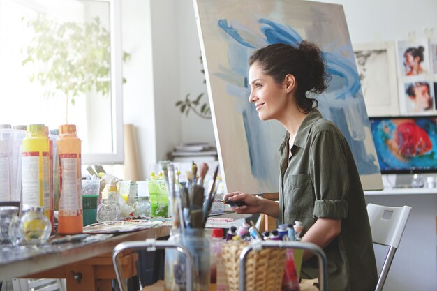 Urocza kobieta ubrana niedbale, patrząc w okno, ciesząc się słońcem pracując w swoim warsztacie, tworząc piękny obraz, malując kolorowymi olejami. Kobieta malarz rysunek na płótnie