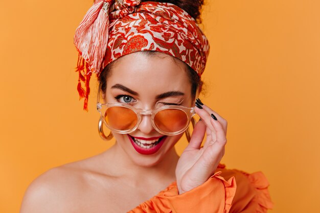 Urocza kobieta w masywnych kolczykach i opasce w stylu afrykańskim zdejmuje pomarańczowe okulary i zalotnie mruga.