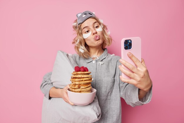 Urocza kobieta wydyma usta i pozuje przed kamerą smartfona pozuje z pysznymi naleśnikami na śniadanie nosi strój do spania trzyma miękką poduszkę nakłada plastry upiększające pozuje na różową ścianę