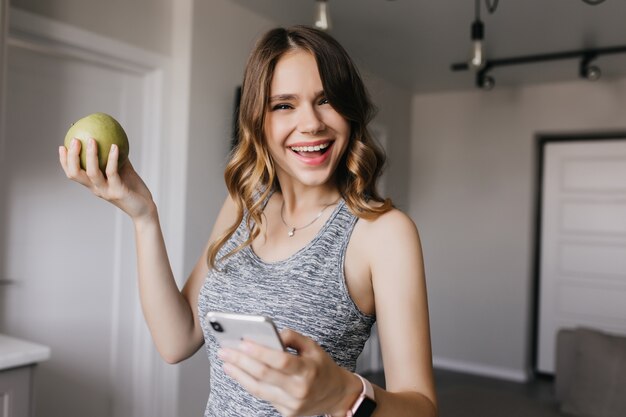 Urocza kobieta z błyszczącymi falującymi włosami, śmiejąca się podczas pozowania w domu. Marzycielska biała dziewczyna trzyma jabłko i smartfon.