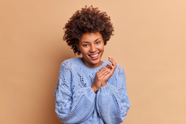 Urocza ładna kobieta ze szczerym uśmiechem naturalne kręcone włosy w stylu afro pocierają dłonie i patrzą radośnie z przodu okazuje radość nosi swobodny sweter odizolowany na beżowej ścianie