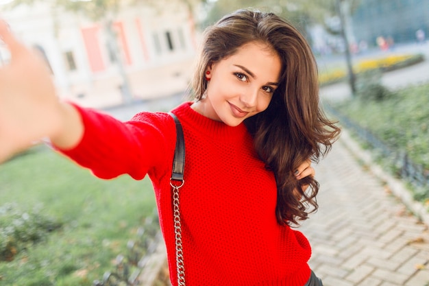 Urocza młoda brunetka kobieta robi autoportret przez telefon komórkowy, spacery w parku, zabawy. Czerwony sweter na co dzień. Styl życia. Świeży makijaż. Falowana fryzura.