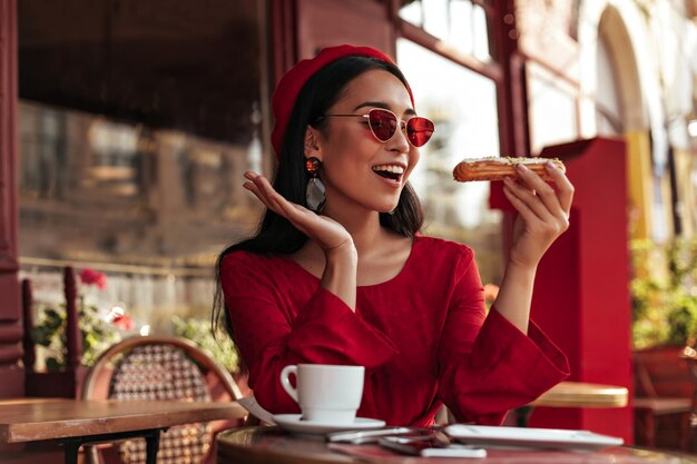 Urocza młoda brunetka w czerwonym berecie w jasnych kolorowych okularach przeciwsłonecznych i stylowej sukience patrzy na smaczną eclair abc w przytulnej ulicznej kawiarni