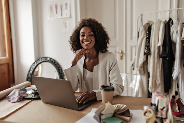 Urocza młoda ciemnoskóra kobieta w stylowej kurtce i bluzce uśmiecha się, patrząc na aparat, który pracuje w laptopie i pozuje w biurze