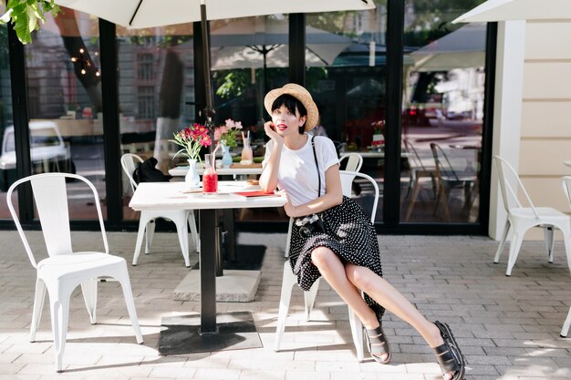 Urocza młoda dama w letnim kapeluszu odpoczywa w kawiarni na świeżym powietrzu, podpierając twarz ręką i oczekującego przyjaciela