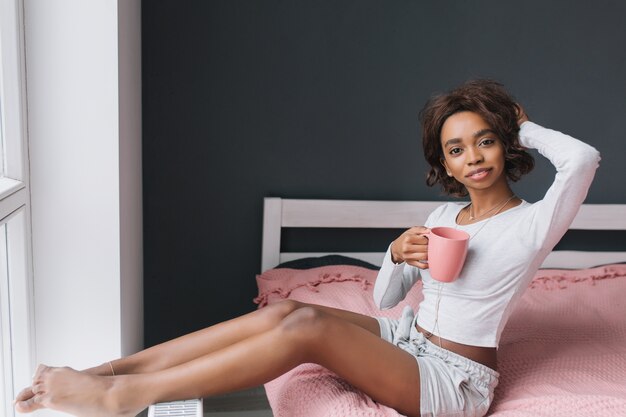 Urocza młoda dziewczyna, ciesząc się rano na łóżku obok okna, trzymając kawę, herbatę w pokoju z szarą ścianą, różowy dywan na przestrzeni