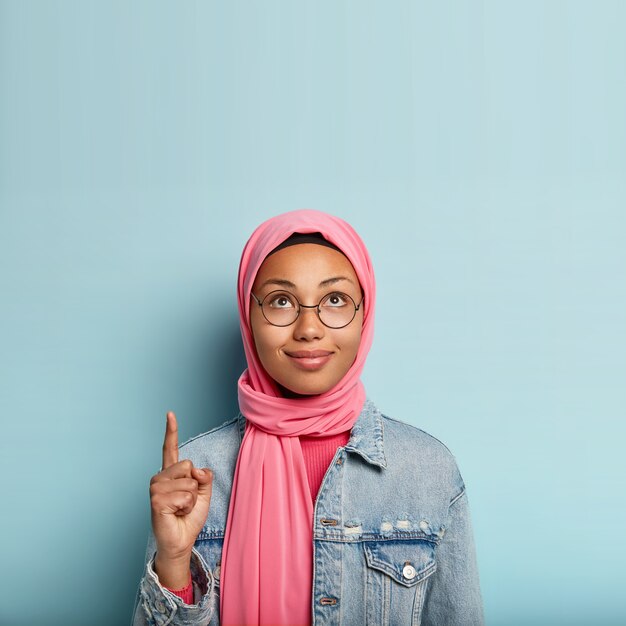 Urocza młoda kobieta o ciemnej, zdrowej skórze, wygląda i wskazuje w górę, wskazuje palcem wskazującym, nosi różowy hidżab, modną kurtkę, stoi nad niebieską ścianą, reklamuje miejsce do kopiowania, wskazuje właściwą drogę