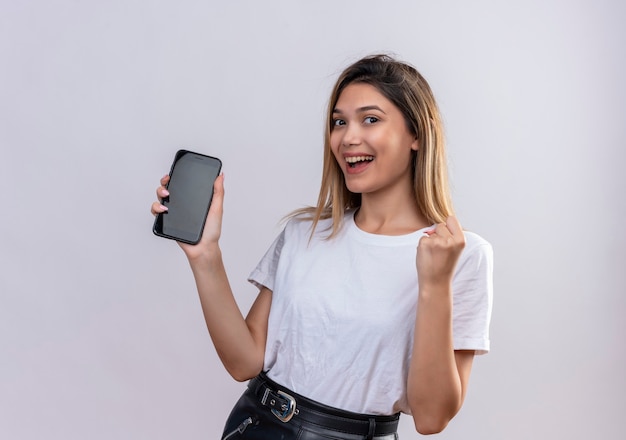 Urocza młoda kobieta w białej koszulce uśmiecha się, pokazując puste miejsce telefonu komórkowego z zaciśniętą pięścią na białej ścianie