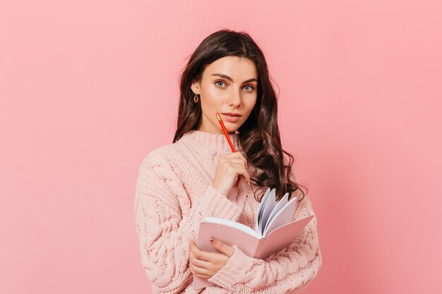 Urocza niebieskooka dziewczyna w zamyśleniu pozuje na różowym tle. Pani z kręconymi włosami trzymająca czerwony ołówek i pamiętnik.