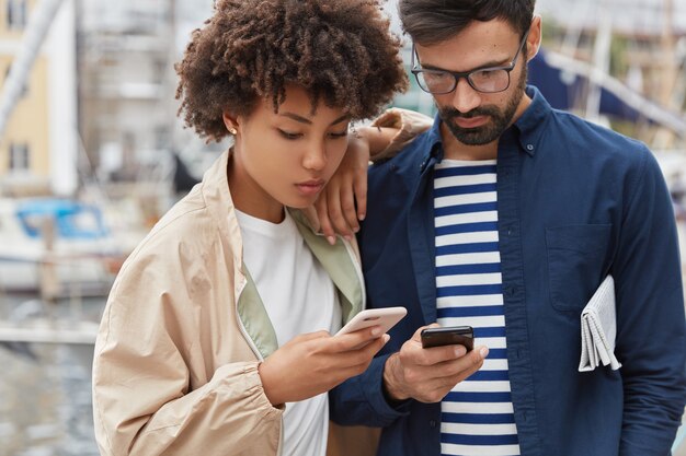 Urocza para stoi obok siebie na niewyraźnym tle zewnątrz, trzyma nowoczesny telefon komórkowy