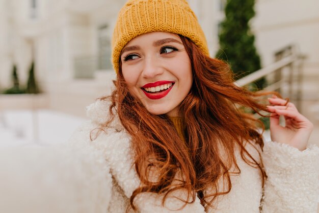 Urocza rudowłosa dziewczyna cieszy się zimą. Plenerowe zdjęcie beztroskiej kaukaskiej pani robiącej selfie w zimny dzień.