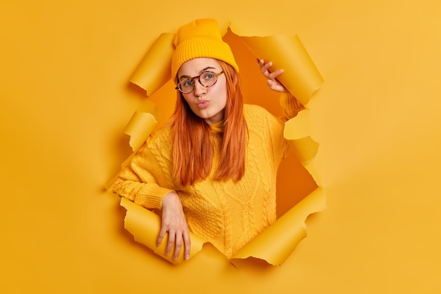 Bezpłatne zdjęcie urocza rudowłosa młoda kobieta z okrągłymi ustami, ubrana w kapelusz i sweter, ma flirt, ubrana w żółte ubranie, stoi przez rozdarty papier
