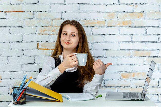 Urocza uczennica siedząca za biurkiem trzymająca filiżankę herbaty i uśmiechnięta Wysokiej jakości zdjęcie