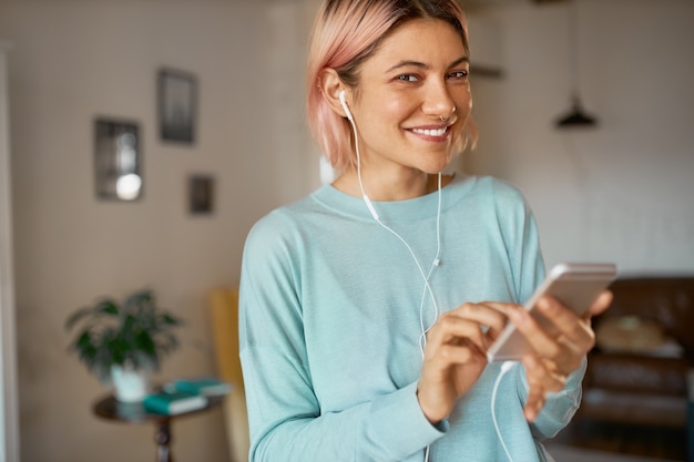 Urocza wesoła młoda kobieta rasy kaukaskiej w słuchawkach trzymając smartfon, ciesząc się wysokiej jakości utworami muzycznymi, pisząc wiadomość tekstową do przyjaciela