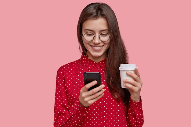 Urocza, zadowolona młoda kobieta trzyma gorącą kawę na wynos, telefon komórkowy, cieszy się, że otrzymuje nowe urządzenie jako prezent, nosi czerwoną koszulę, ma delikatny uśmiech