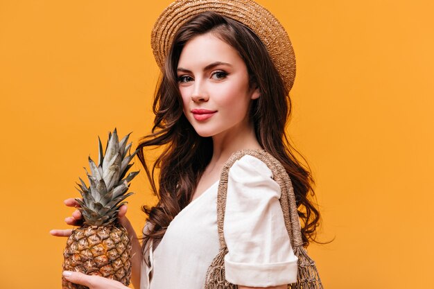 Urocza zielonooka brunetka kobieta w słomkowym kapeluszu, trzymając ananas i patrząc w kamerę na pomarańczowym tle.