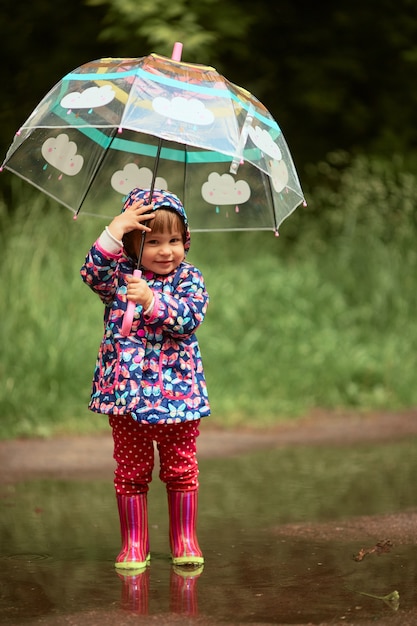 Urocze dziewczynki z parasolem ma zabawy stojące w gumboots w basenie po deszczu