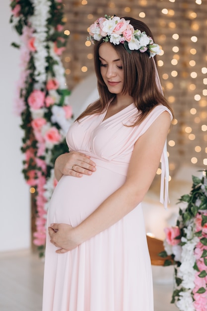 Uroczy brunetka kobieta w ciąży w różowej sukience pozuje w Wieniec kwiatów