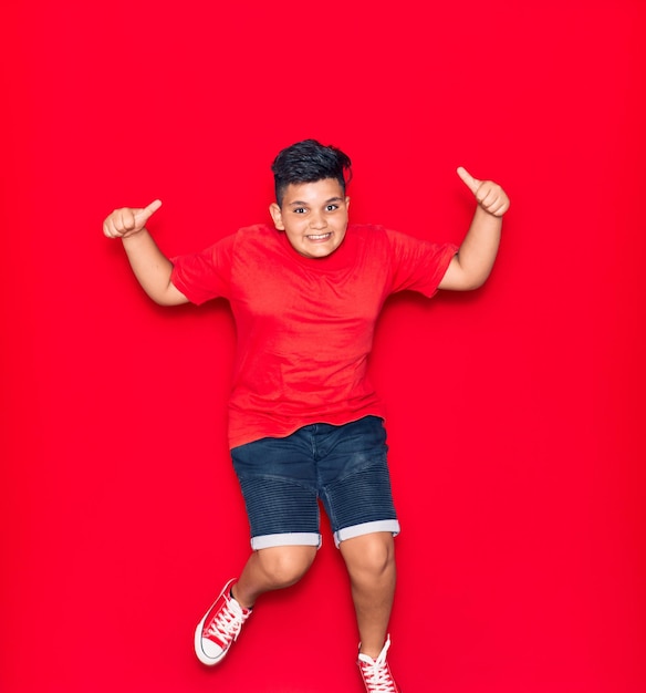 Bezpłatne zdjęcie uroczy dzieciak ubrany w przypadkowe ubrania uśmiechnięty szczęśliwy skakanie z uśmiechem na twarzy robi znak ok z aprobatami na odosobnionym czerwonym tle