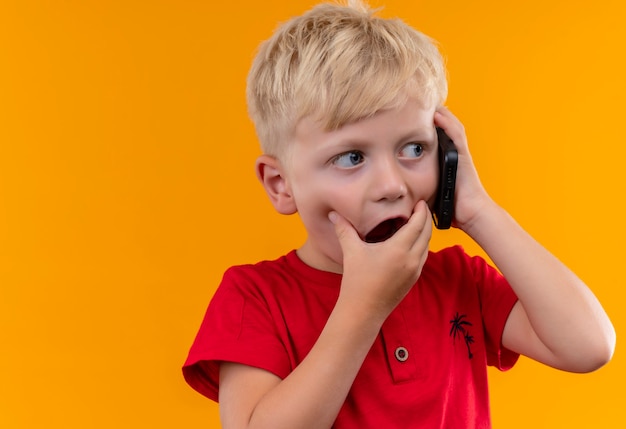 Uroczy mały chłopiec o blond włosach i niebieskich oczach w czerwonej koszulce mówiący przez telefon komórkowy, patrząc zaskakująco z boku z ręką na ustach