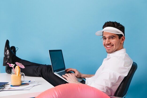 Uroczy mężczyzna w białej koszuli, czapce i okularach jest na tle niebieskiej przestrzeni. Facet trzyma laptopa i pracuje na wakacjach.