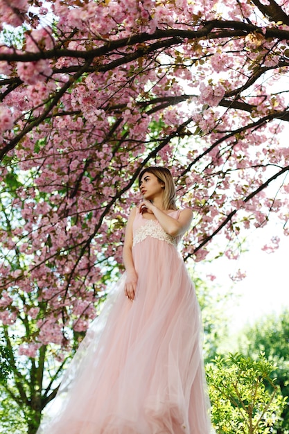 Uroczy młoda kobieta w różowej sukience stawia przed sakura drzewa pełne różowe kwiaty