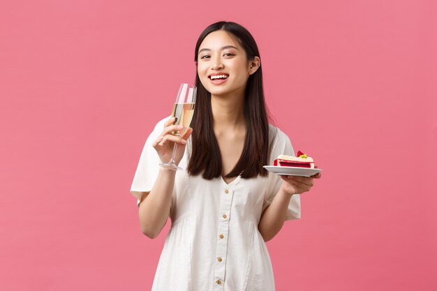 Uroczystości, święta i zabawy koncepcja. Happy asian kobieta świętuje urodziny, ciesząc się smacznym tortem urodzinowym i pijąc szampana, uśmiechając się do kamery radosne, różowe tło.
