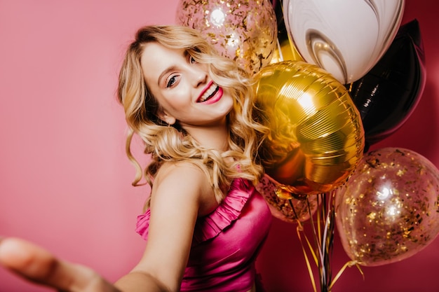 Bezpłatne zdjęcie urodzinowa dziewczyna debonair robi selfie wewnątrz zdjęcie wyrafinowanej blondynki damy pozującej na różowym tle z balonami