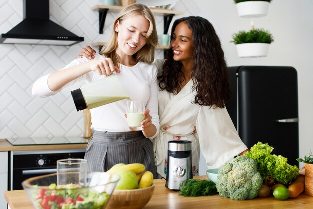 Uśmiechająca się atrakcyjna oliwkowa kobieta w bieliźnie nocnej i kaukaska ze zdrowym smoothie stoi przy stole pełnym świeżych owoców i warzyw na białej nowoczesnej kuchni