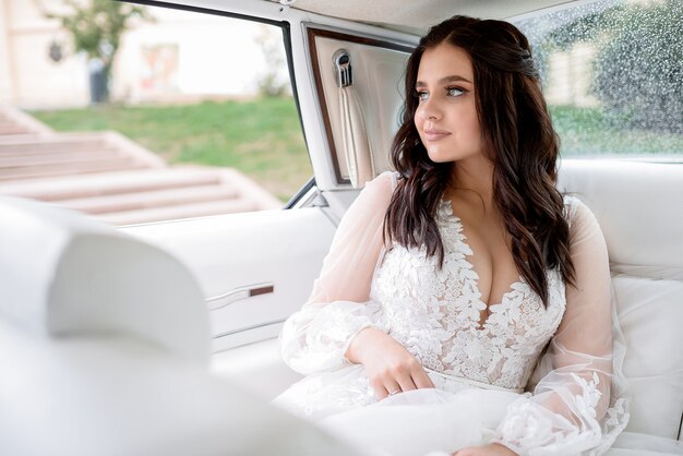 Uśmiechająca się piękna panna młoda brunetka z otwartym dekoltem siedzi w samochodzie i patrzy przez okno