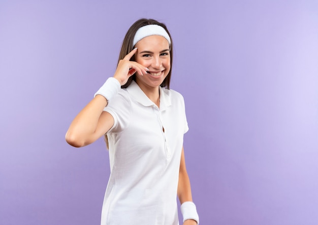 Uśmiechający się całkiem sportowy dziewczyna nosi opaskę i opaskę kładąc palec na świątyni na białym tle na fioletowej przestrzeni