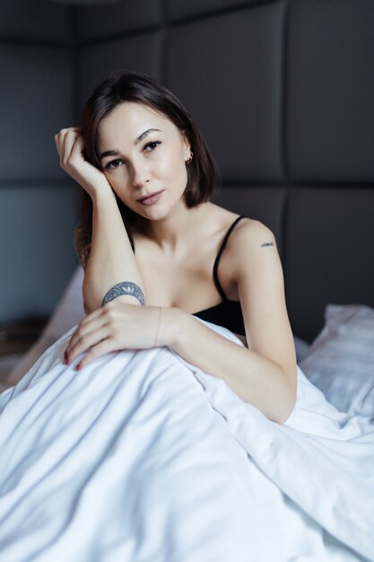 Uśmiechający się Długie włosy brunetka kobieta na białym łóżku w miękkim świetle poranka pod kołdrą