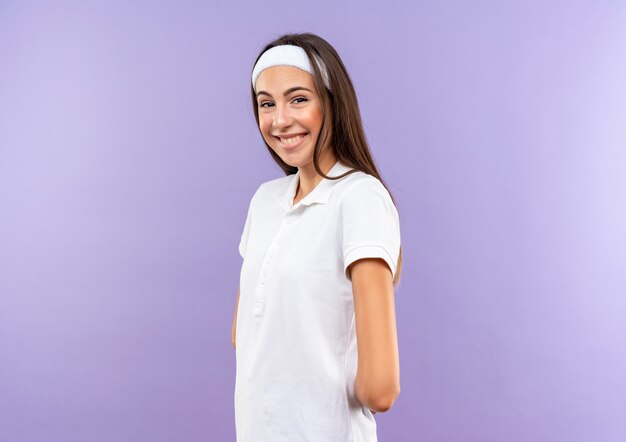 Uśmiechający się ładny sportowy dziewczyna noszenie opaski i opaski stojącej w widoku profilu z rękami za plecami na białym tle na fioletowej przestrzeni