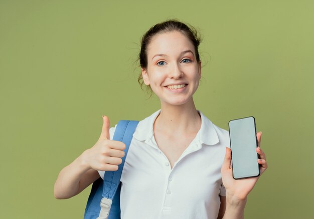 Uśmiechający się młody ładny studentka noszenie plecaka pokazujący telefon komórkowy i kciuk na białym tle na zielonym tle