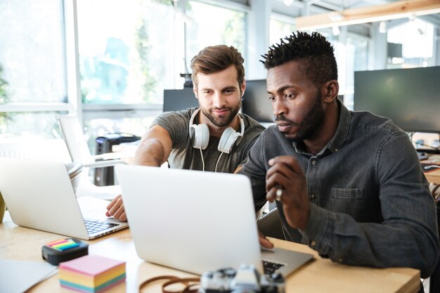 Uśmiechnięci młodzi koledzy siedzi w biurowym coworking używać laptop