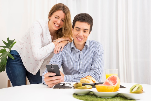 Bezpłatne zdjęcie uśmiechnięci potomstwa dobierają się patrzeć telefon komórkowego przy śniadaniowym stołem