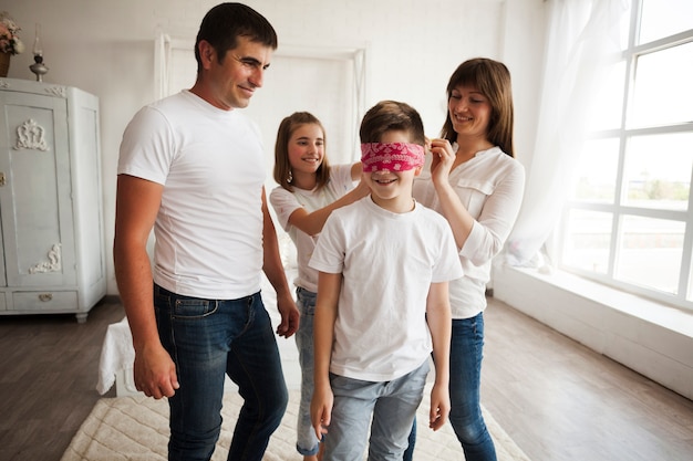 Uśmiechnięci rodzice patrząc na swoją córkę wiążącą szalik w oczach brata
