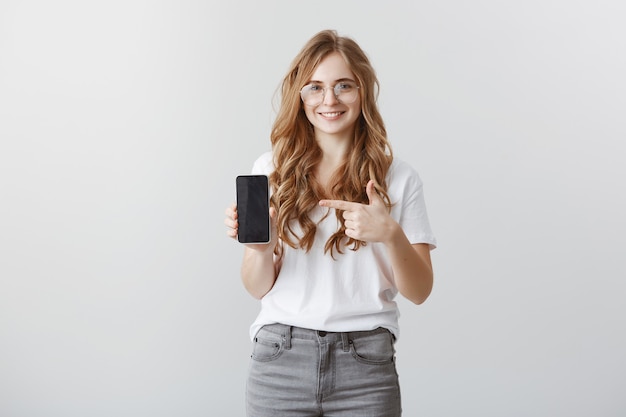 Bezpłatne zdjęcie uśmiechnięta atrakcyjna blond dziewczyna w okularach wskazując palcem na ekranie smartfona, pokazując aplikację