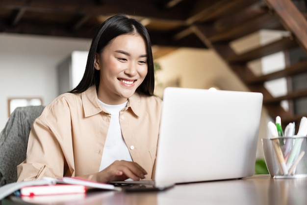 Uśmiechnięta Azjatka oglądająca webinarium, prowadząca rozmowę wideo z domu, pracująca jako freelancer, zdal...