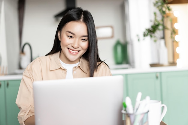 Uśmiechnięta Azjatka używa laptopa pracującego w domu na komputerze PC, siedząc w kuchni i studiując...