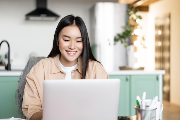 Uśmiechnięta Azjatka używa laptopa pracującego w domu, ucząc się w szkole online na zdalnym uniwersytecie...